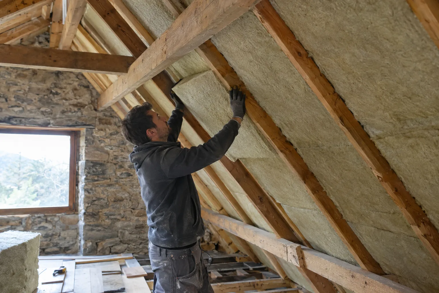 Couvreur qui déroule un isolant entre les chevrons dans les combles d'une maison d'Aubagne, tuiles déposées et lumière entrant par la toiture ouverte.