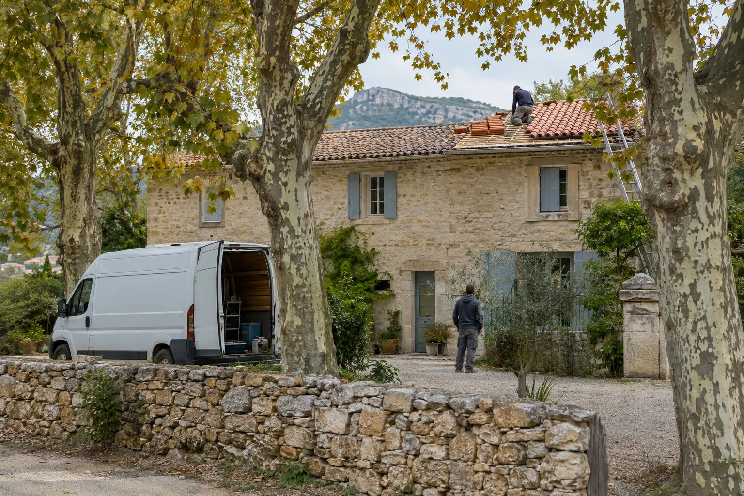 Couvreur sur le toit d’une grande bastide en pierre entourée de platanes, camionnette ouverte devant la maison, colline en arrière-plan